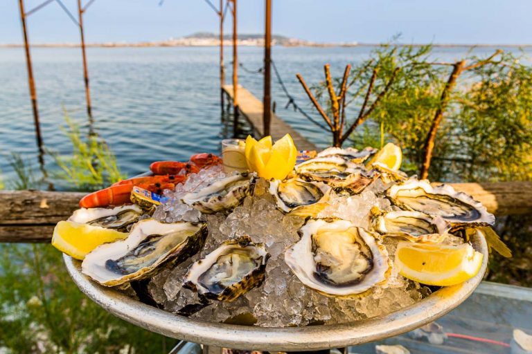 Plato de ostras con marisco en el pabellón de playa Le St Pierre Tarbouriech. Detrás de la laguna se encuentra la ciudad de Sète / © Foto: Georg Berg